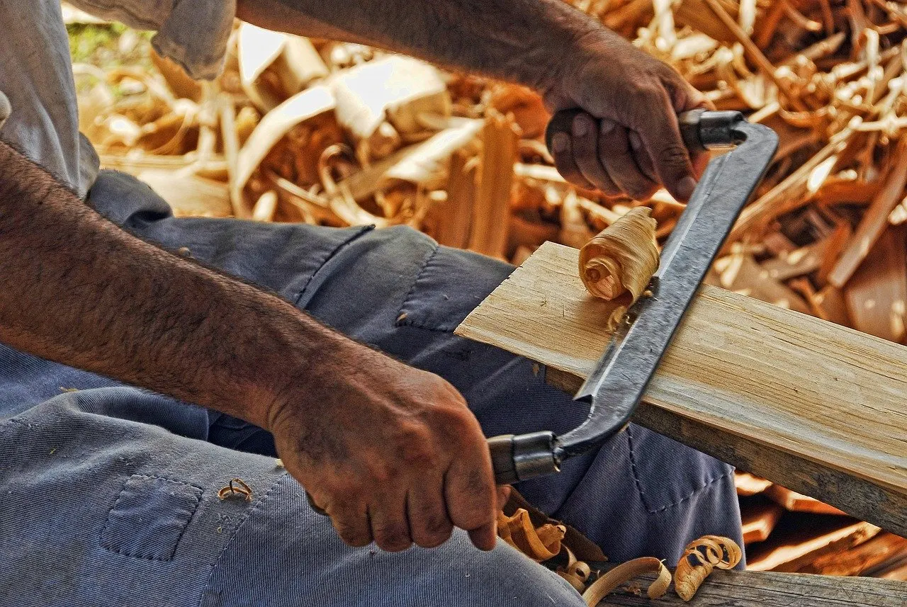 Zwei Hände führen ein Zugmesser über ein helles Holzbrett, um das Brett herum liegen viele gekräuselte Holzspäne