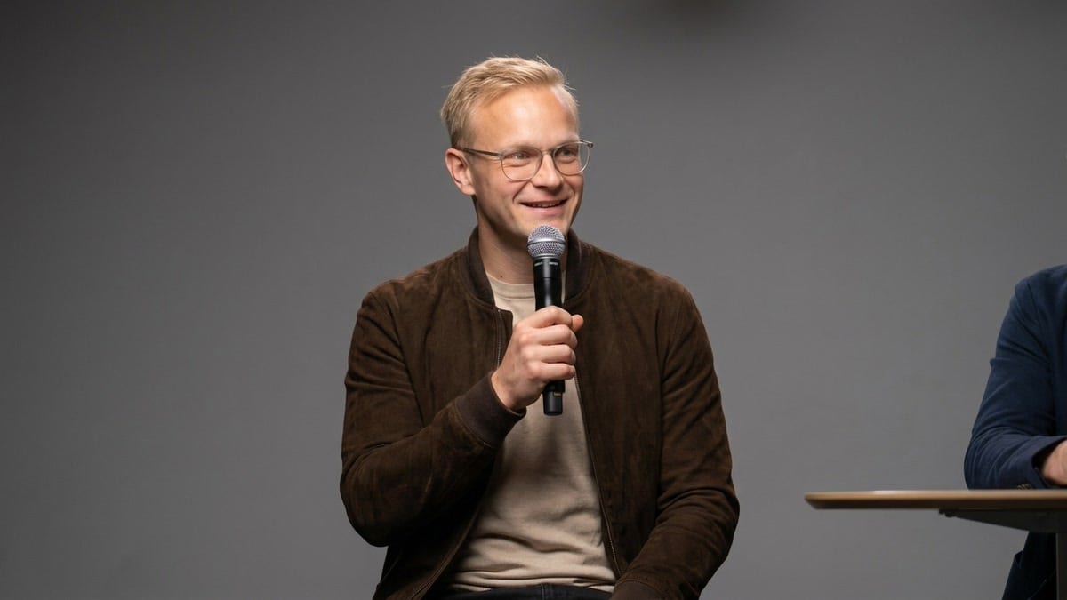 Sebastian Wagner sitzt mit Mikrofon auf einem Hocker und spricht lächelnd vor grauem Hintergrund