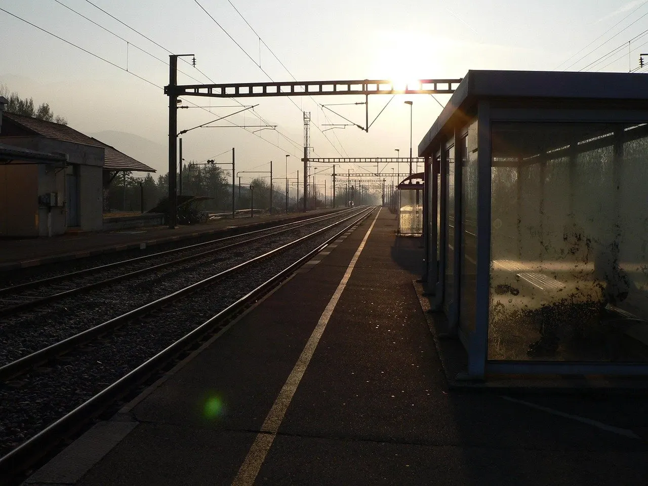 Leerer Bahnsteig im Gegenlicht der Morgensonne, links mehrere Gleise mit Oberleitung, rechts ein verglastes Wartehäuschen