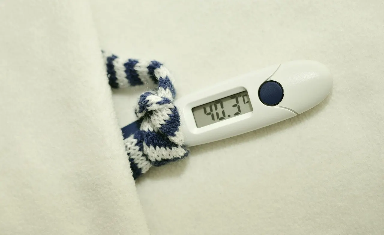 Digitales Fieberthermometer mit der Anzeige 40,3 Grad auf einer hellen Wolldecke, daneben ein blau-weiß gestreifter Strickschal