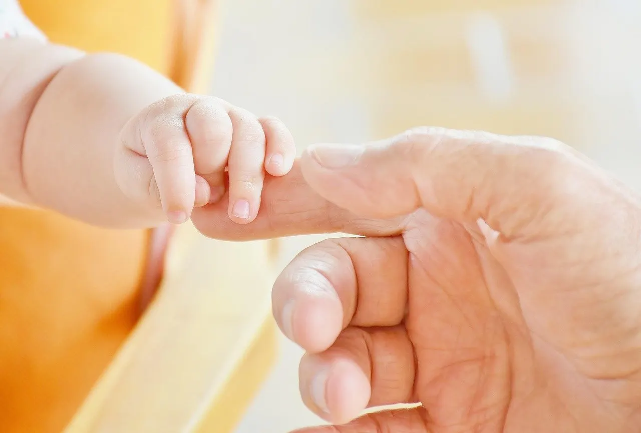 Kleine Babyhand umgreift den Zeigefinger einer Erwachsenenhand, im unscharfen Hintergrund ein gelber Stoff