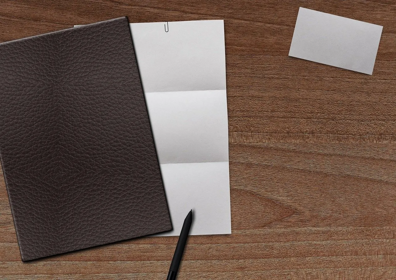 Dunkle Ledermappe neben einem gefalteten weißen Blatt Papier, einer leeren Visitenkarte und einem Stift auf einem Holztisch