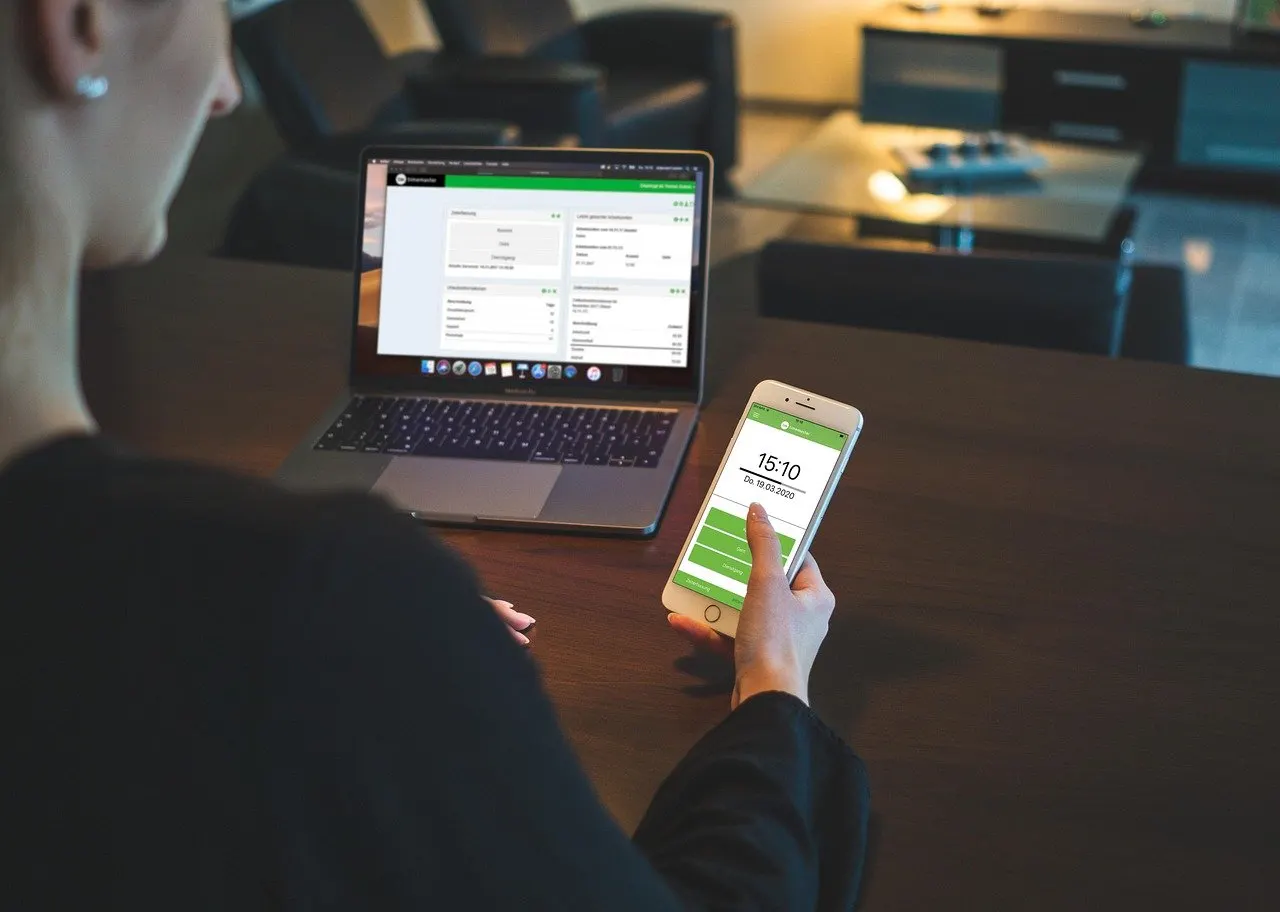 Person am Schreibtisch hält ein Smartphone mit Zeiterfassungs-App und Uhrzeit 15:10, dahinter ein aufgeklappter Laptop