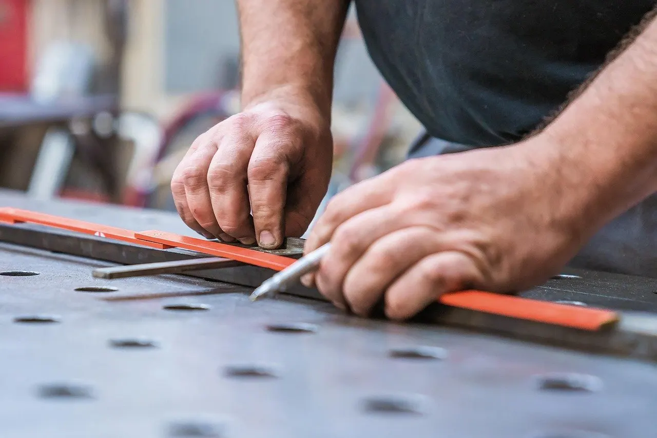 Zwei Hände legen einen orangefarbenen Winkel an ein Metallprofil und reißen mit einem Stift auf einem gelochten Werktisch an