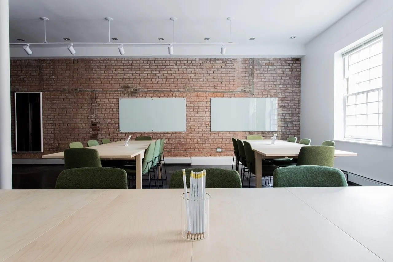 Leerer Schulungsraum mit Backsteinwand, zwei Whiteboards, hellen Tischen und grünen Stühlen, vorne ein Glas mit Bleistiften