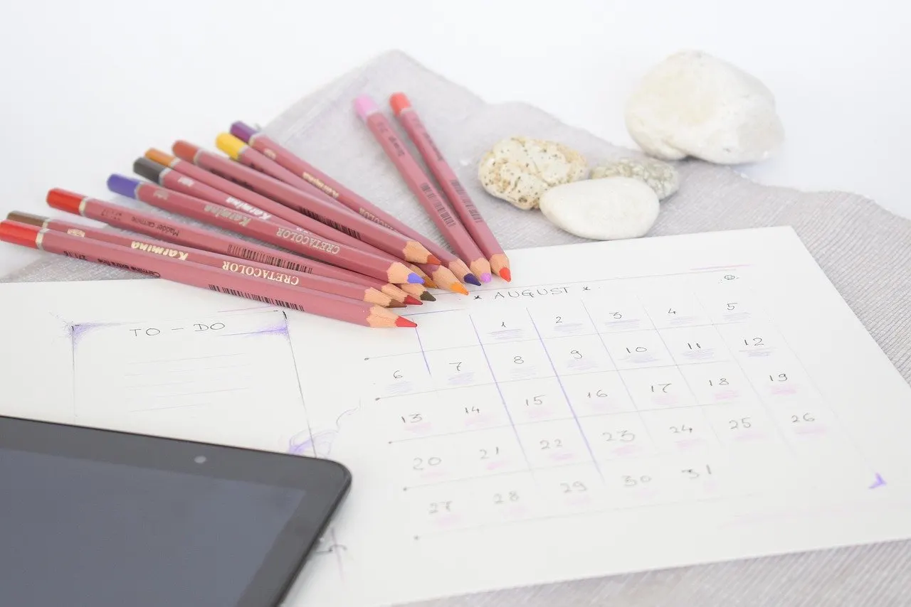 Handgezeichneter Monatskalender für August auf weißem Papier, daneben rosafarbene Buntstifte mit bunten Minen, Kieselsteine und ein Tablet