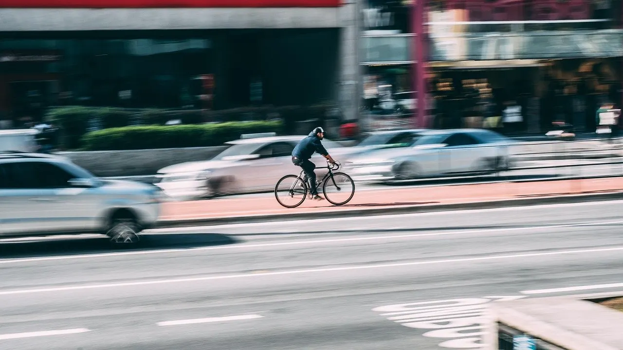 Radfahrer mit Kappe auf einem Radweg an einer mehrspurigen Stadtstraße, daneben unscharf vorbeifahrende Autos im Berufsverkehr